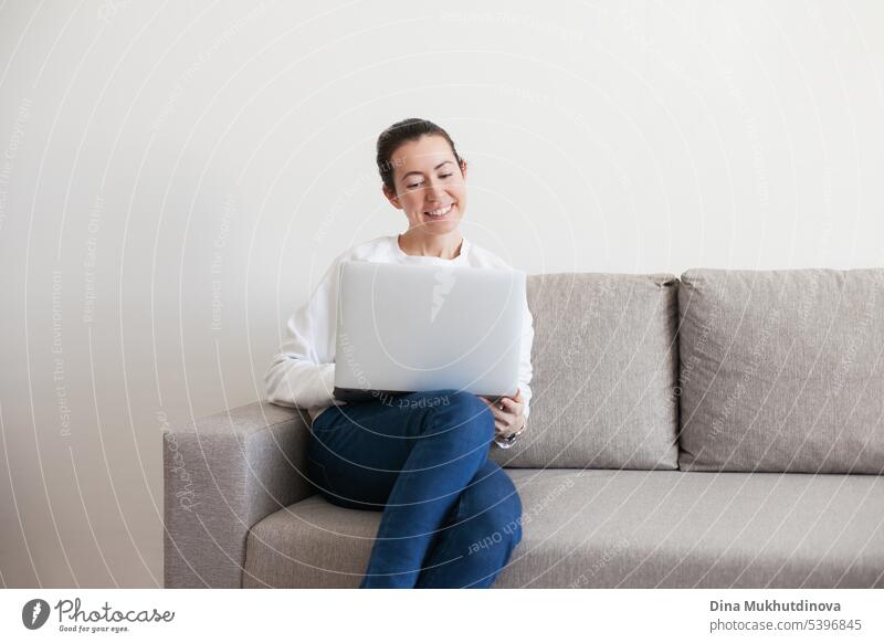 Junge Frau arbeitet am Laptop. Millennial Mädchen arbeiten Remote-Job von zu Hause aus, sitzen auf dem gemütlichen Sofa, lächelnd. Hintergrund schön Business