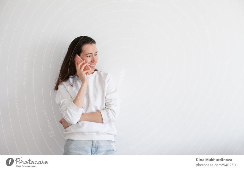 Junge Frau hält ein Mobiltelefon, lächelnd, mit sozialen Medien. Millennial Person in weißem Sweatshirt auf weißem Hintergrund. Menschen nutzen Technologie. Anruf bei der Arbeit.