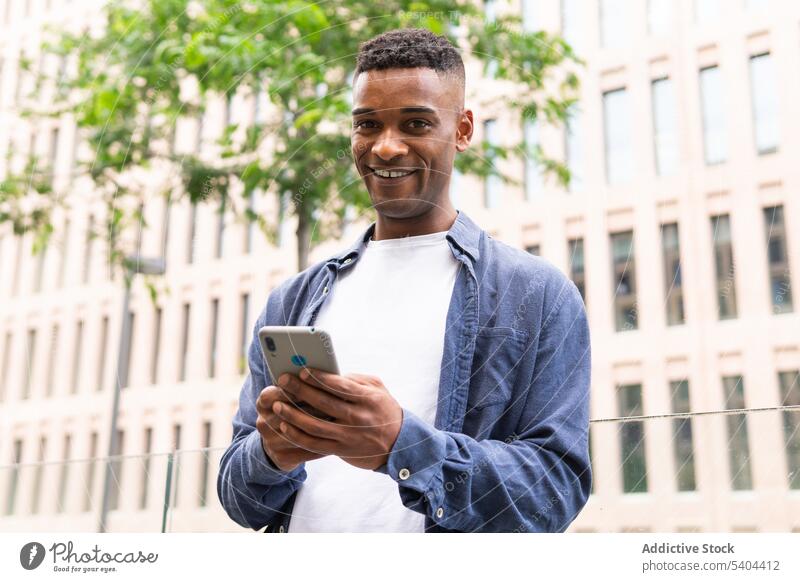 Positiver schwarzer Mann, der mit seinem Smartphone chattet Unternehmer Mobile Textnachricht Talkrunde Arbeit Lächeln Straße Großstadt männlich Afroamerikaner