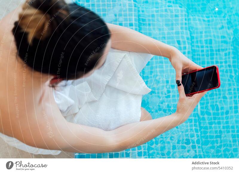 Frau in weißem Handtuch sitzt am Pool und surft auf ihrem Smartphone Urlaub benutzend Beckenrand ruhen Browsen Gerät Bademantel Resort Hotel