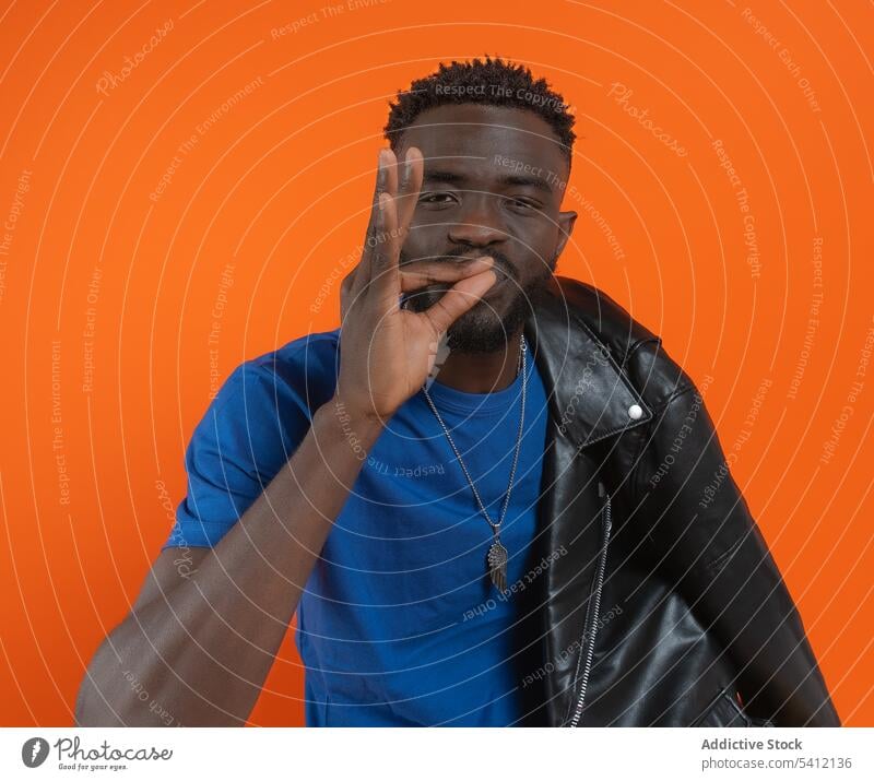 Seriöser junger schwarzer Mann sitzt und zeigt OK-Zeichen in der Nähe der orangefarbenen Wand gestikulieren selbstbewusst Porträt ernst selbstsicher Lederjacke