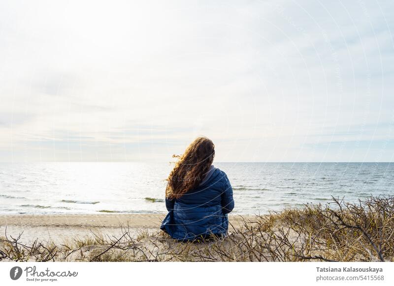 Langhaarige lockige Plus-Size-Frau am Meeresufer, Rückansicht Lockenhaarige Übergröße dünne Daunenjacke Ostsee Abend Wind Behaarung flatternd Natur Schönheit