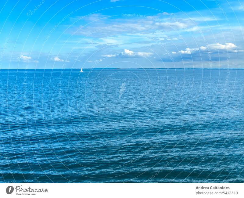 Blaues Bild, zwei Drittel Ostsee, ein Drittel leicht bewölkter Himmel, links oben ein kleines einzelnes weißes Segelboot, im Vordergrund leichte Wellen, horizontal