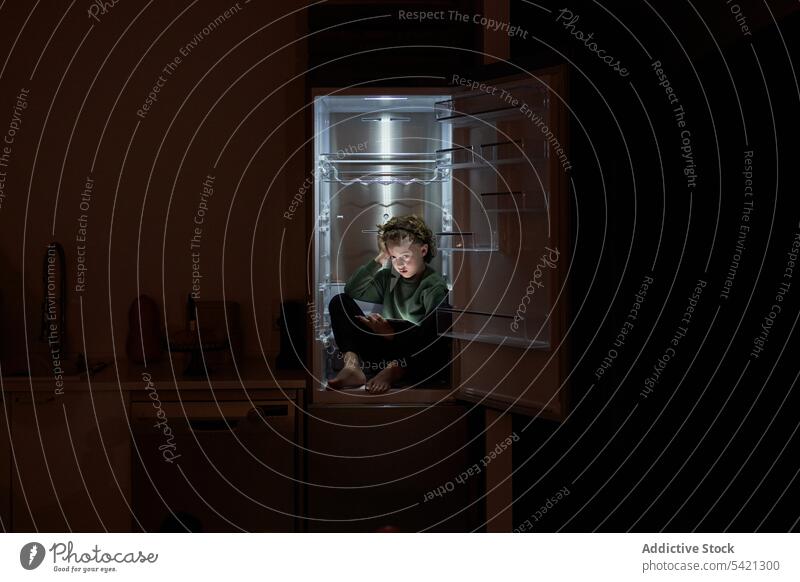 Ruhiges, entspanntes Kind, das in einem leeren Kühlschrank sitzt und in einer heißen Nacht ein digitales Tablet benutzt Tablette essen sich[Akk] entspannen