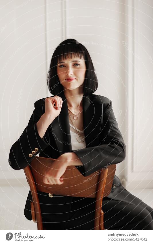 attraktive Business-Frau sitzt auf einem Holzstuhl und schaut in die Kamera. brünette Dame mit kurzen Haaren trägt schwarzen Büroanzug und weiße Bluse, High-Heels auf weißem Wandhintergrund.
