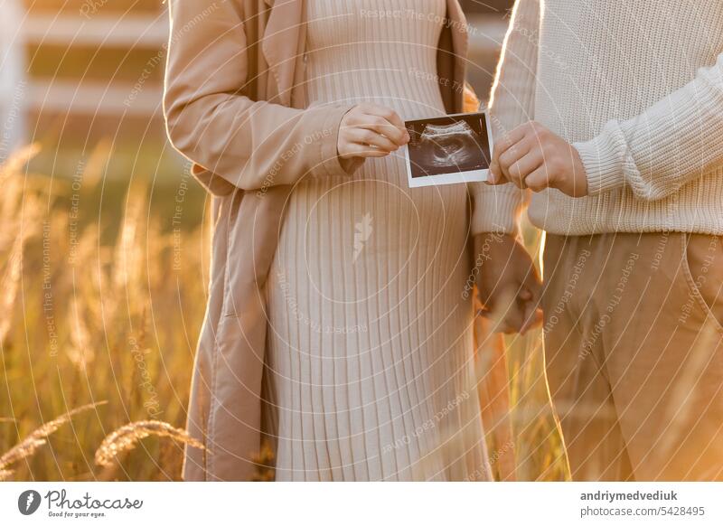 Unerkannt von einer schwangeren Frau mit ihrem Mann halten ein Ultraschall-Scan-Foto des ungeborenen Kindes im Freien auf die Natur in Sonnenlicht. Glückliche Mutterschaft und Elternschaft Konzept