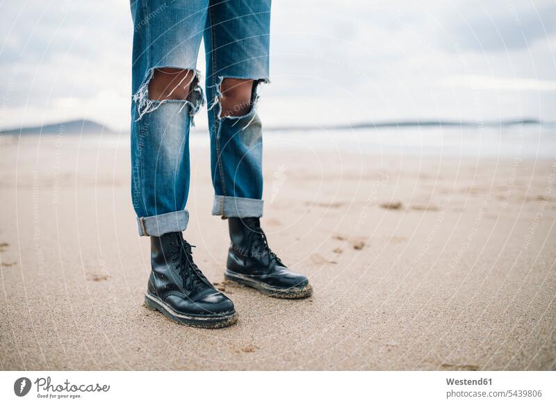 Frau mit Stiefeln und zerrissenen Jeans am Strand, Teilansicht Beach Straende Strände Beaches weiblich Frauen zerfetzt gerissen Jeanshose stehen stehend steht