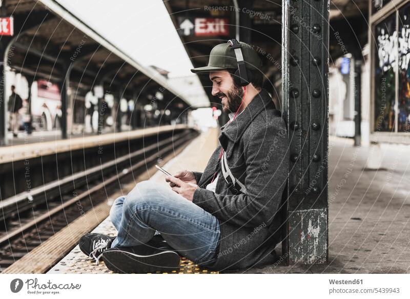Junger Mann wartet am Bahnsteig auf die U-Bahn und benutzt ein Smartphone Handy Mobiltelefon Handies Handys Mobiltelefone warten Musik Kopfhörer Kopfhoerer