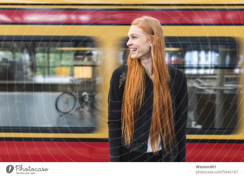 Lachende rothaarige junge Frau wartet am Bahnsteig, Berlin, Deutschland freuen zufrieden stehend steht Muße Lifestyles mobil Spass spassig spaßig Spässe Späße