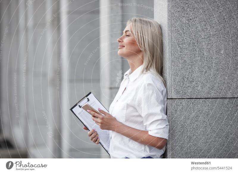 Lächelnde Geschäftsfrau mit Klemmbrett und an die Wand gelehntem Handy Geschäftsfrauen Businesswomen Businessfrauen Businesswoman Mobiltelefon Handies Handys