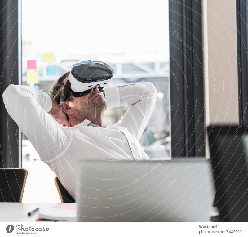Geschäftsmann mit VR-Brille lehnt sich am Schreibtisch im Büro zurück Businessmann Businessmänner Geschäftsmänner Office Büros Geschäftsleute Geschäftspersonen