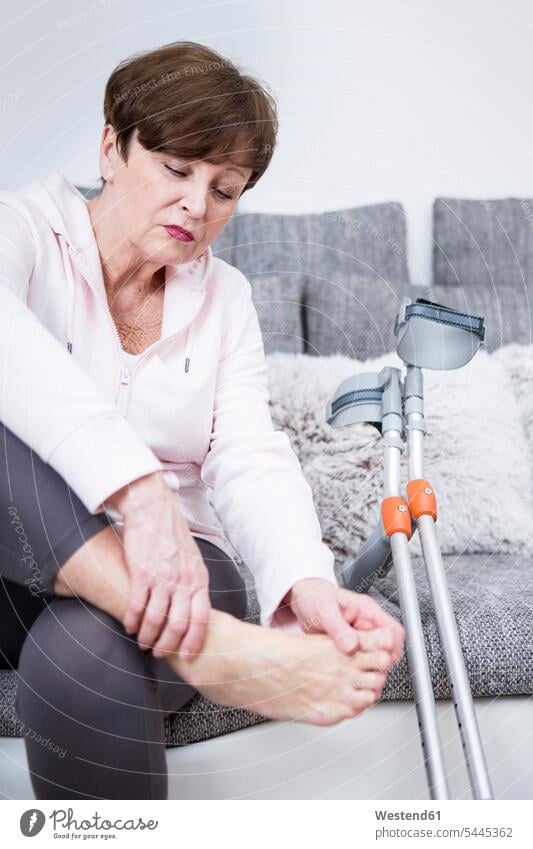 Ältere Frau mit Krücken, die auf einer Couch sitzt und ihren Knöchel kontrolliert betrachten betrachtend Kruecken Fuß Fuss Füße Schmerz Schmerzen Fußknöchel