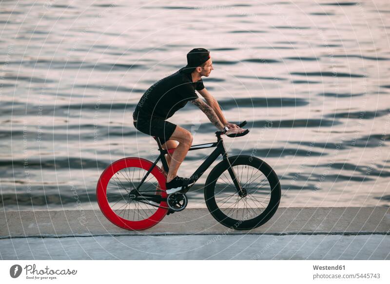 Junger Mann fährt Fixie-Rad am Wasser fahren fahrend fahrender fahrendes Fahrrad Bikes Fahrräder Räder Männer männlich Fluss Fluesse Fluß Flüsse Raeder