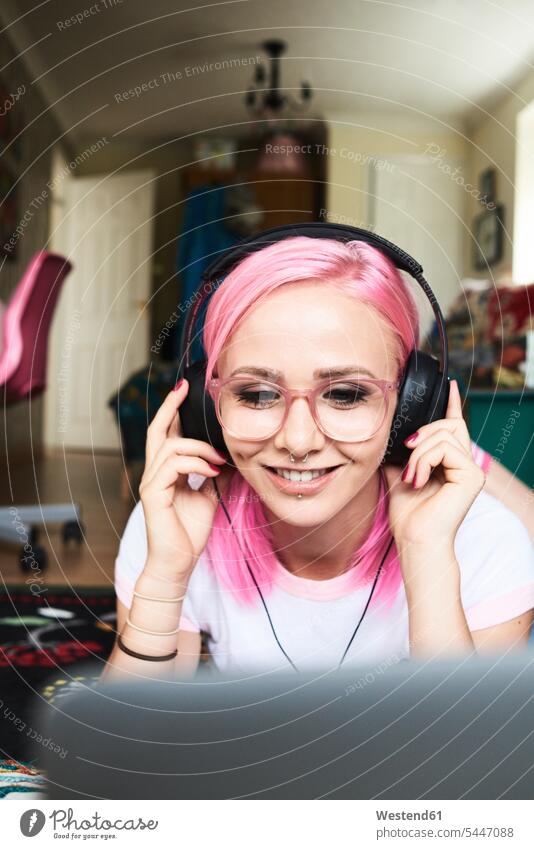 Junge Frau mit rosa Haaren hört zu Hause Musik über ihren Laptop weiblich Frauen Kopfhörer Kopfhoerer Brille Brillen fröhlich Fröhlichkeit Frohsinn Heiterkeit