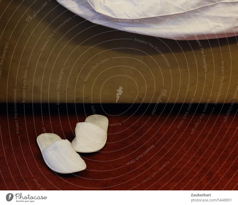 Weiße Pantoffeln stehen vor einem Hotelbett, darüber das Bettlaken Puschen latschen schlappen weiß Teppich Laken schlafen Morgen Abend morgens Schlafzimmer