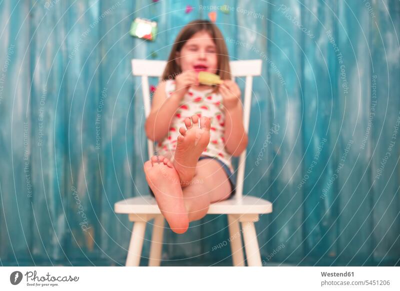 Füße eines kleinen Mädchens, das auf einem Stuhl sitzt und Zitroneneis-Lolly isst Stuehle Stühle Eis Speiseeis weiblich essen essend sitzen sitzend Fuß Fuss