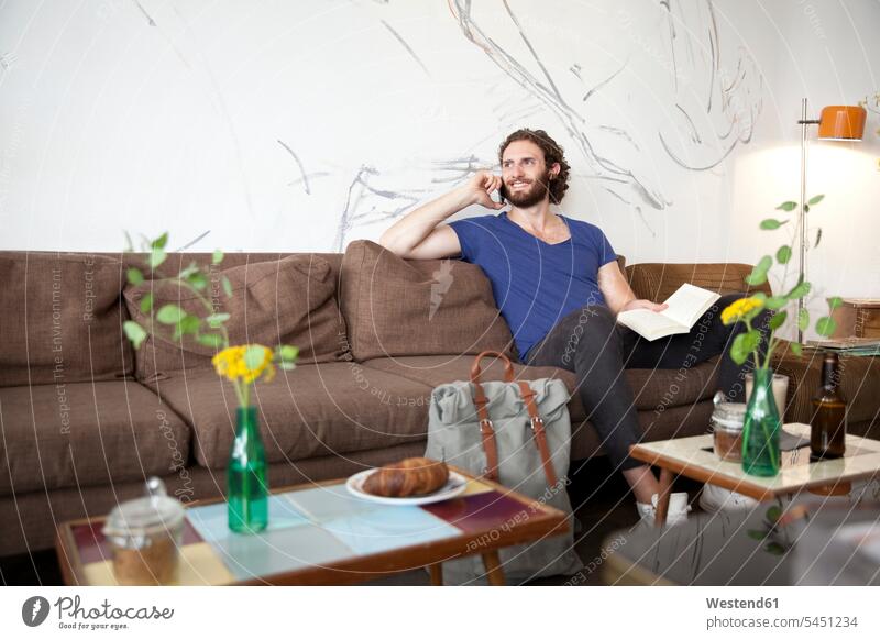 Porträt eines jungen Mannes am Telefon, der in einem Café mit Buch auf der Couch sitzt Cafe Kaffeehaus Bistro Cafes Kaffeehäuser telefonieren anrufen Anruf