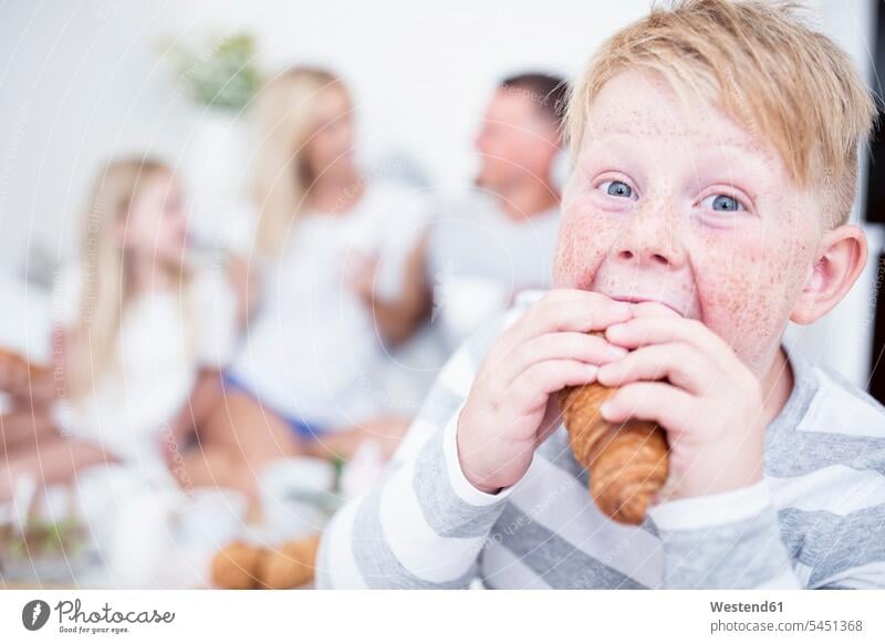 Porträt eines in ein Croissant beißenden Jungen mit Familie im Hintergrund Hoernchen Croissants Cornetto Cornettos Hörnchen Frühstück frühstücken essen essend