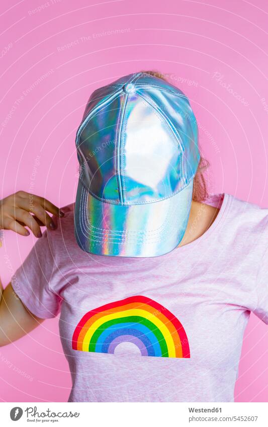 Junge Frau mit Kopfhörern versteckt ihr Gesicht hinter einer Basecap vor rosa Hintergrund Schirmmuetze Mütze Schirmmütze Muetze Kopfbedeckung Baseballmütze