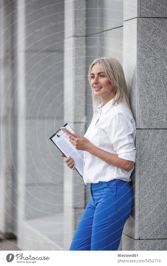 Lächelnde Geschäftsfrau mit Klemmbrett und an die Wand gelehntem Handy lächeln Geschäftsfrauen Businesswomen Businessfrauen Businesswoman Mobiltelefon Handies