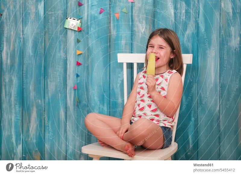 Glückliches kleines Mädchen sitzt auf einem Stuhl und isst Zitroneneis-Lolly sitzen sitzend Stuehle Stühle Eis Speiseeis essen essend weiblich Süßspeise Süsses