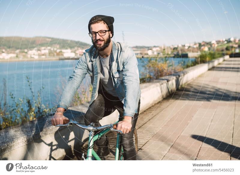 Lächelnder junger Mann auf seinem Fixie-Bike am Wasser Männer männlich lächeln fahren fahrend fahrender fahrendes Fahrrad Bikes Fahrräder Räder Rad Erwachsener
