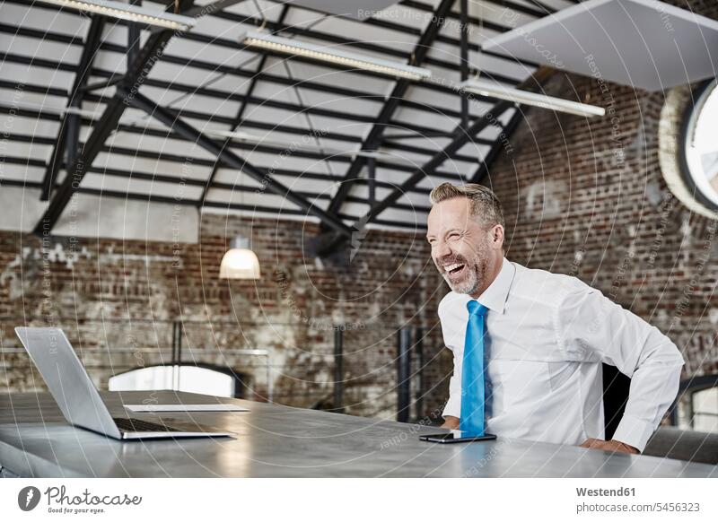 Lachender Geschäftsmann mit Laptop sitzt am Tisch in einem Loft sitzen sitzend Notebook Laptops Notebooks lachen Businessmann Businessmänner Geschäftsmänner