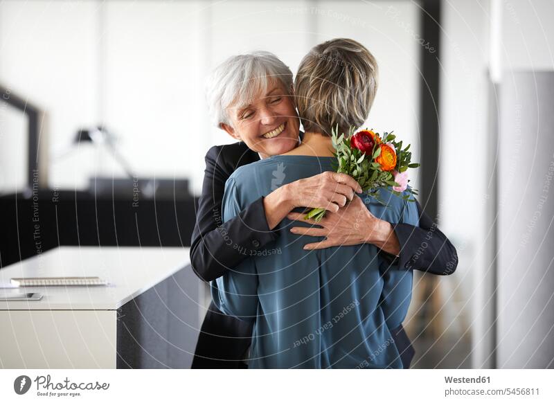 Glückliche ältere Geschäftsfrau mit Blumenstrauss umarmt Amtskollegin Generation Arbeitskollege Arbeitskollegen Kollege Job Berufe Berufstätigkeit Beschäftigung