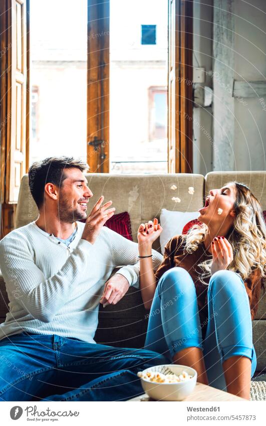 Fröhliches junges Paar sitzt zu Hause und spielt mit Popcorn Couch Couches Liege Sofas sitzend freuen Frohsinn Fröhlichkeit Heiterkeit geniessen Genuss Glück