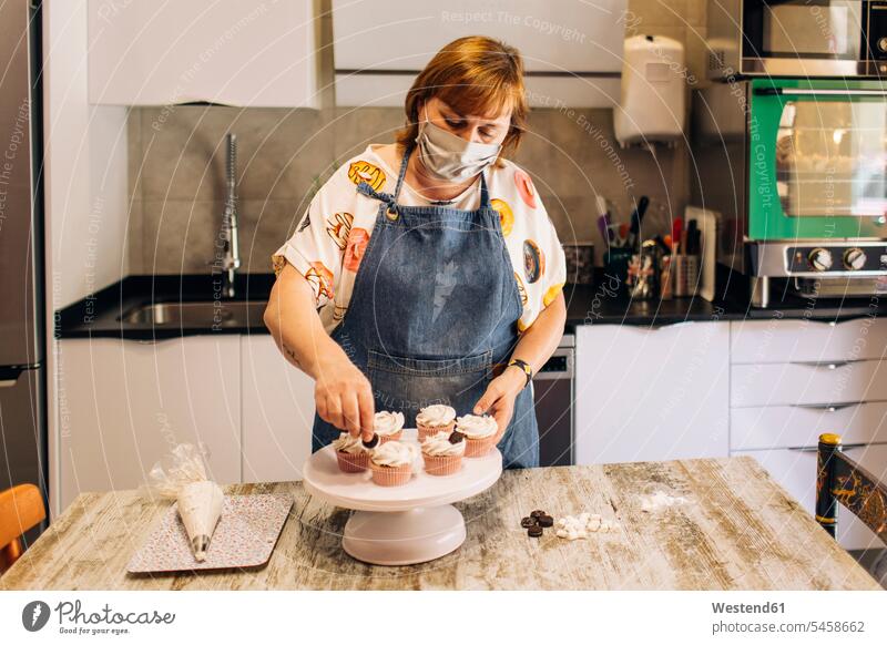 Bäckerin mit Maske, die Törtchen auf Kuchenständer in Werkstatt dekoriert Farbaufnahme Farbe Farbfoto Farbphoto Innenaufnahme Innenaufnahmen innen drinnen
