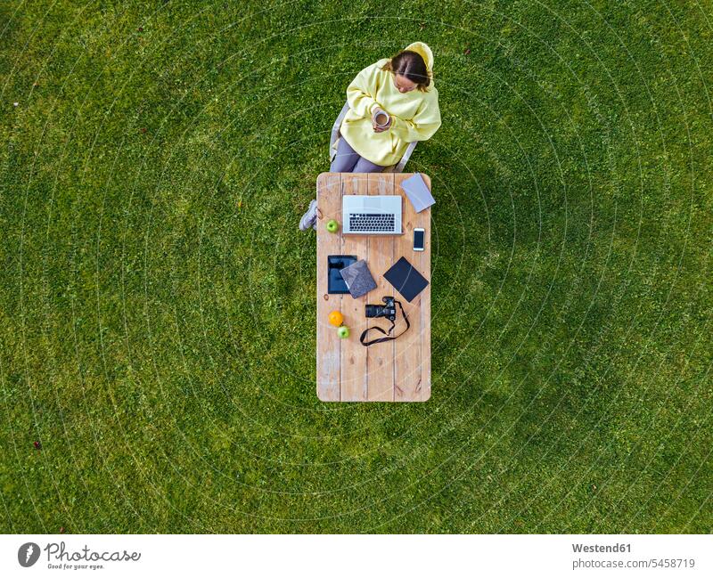 Luftaufnahme einer Frau an einem auf grünem Rasen gedeckten Kaffeetisch mit einem Becher Kaffee in der Hand Außenaufnahme außen draußen im Freien Tag
