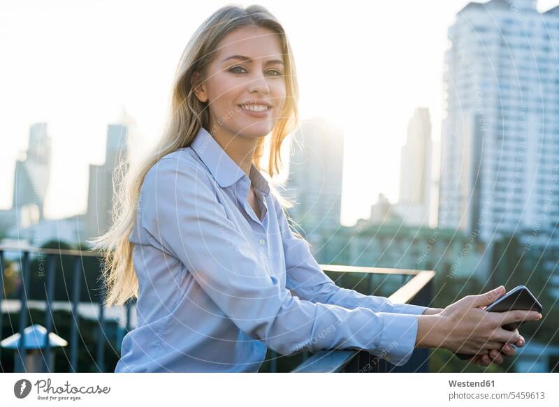 Blonde lächelnde Geschäftsfrau lehnt sich an ein Geländer und hält ein Smartphone auf dem Dach der Stadt Geschäftsfrauen Businesswomen Businessfrauen