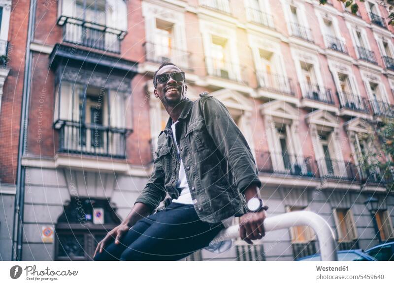 Lächelnder Mann mit Sonnenbrille sitzt gegen Gebäude in der Stadt Farbaufnahme Farbe Farbfoto Farbphoto Spanien Freizeitbeschäftigung Muße Zeit Zeit haben
