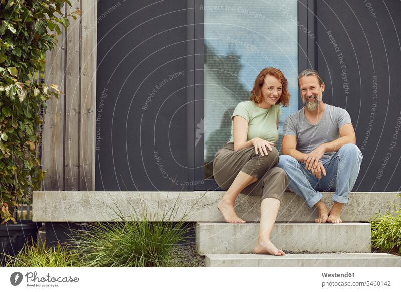 Lächelndes Paar sitzt auf Stufen vor winzigem Haus Farbaufnahme Farbe Farbfoto Farbphoto Deutschland Freizeitbeschäftigung Muße Zeit Zeit haben Freizeitkleidung