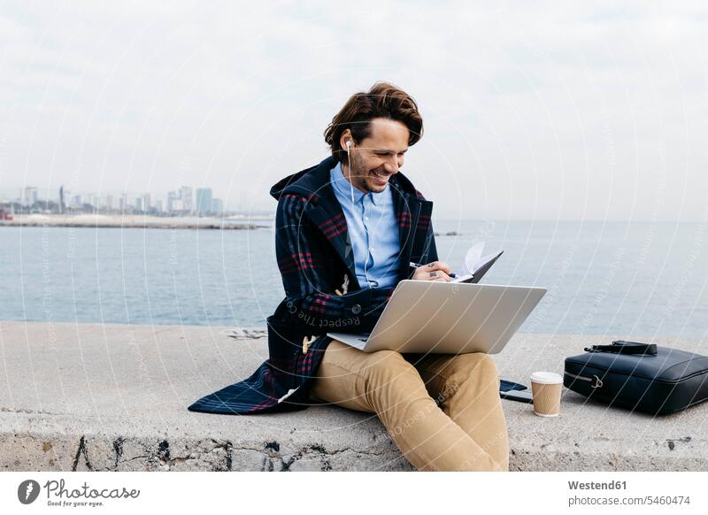 Spanien, Barcelona, lächelnder Mann sitzt am Meer und arbeitet mit Laptop und Notebook sitzen sitzend Männer männlich Notizbuch Notizbücher Notizbuecher Meere