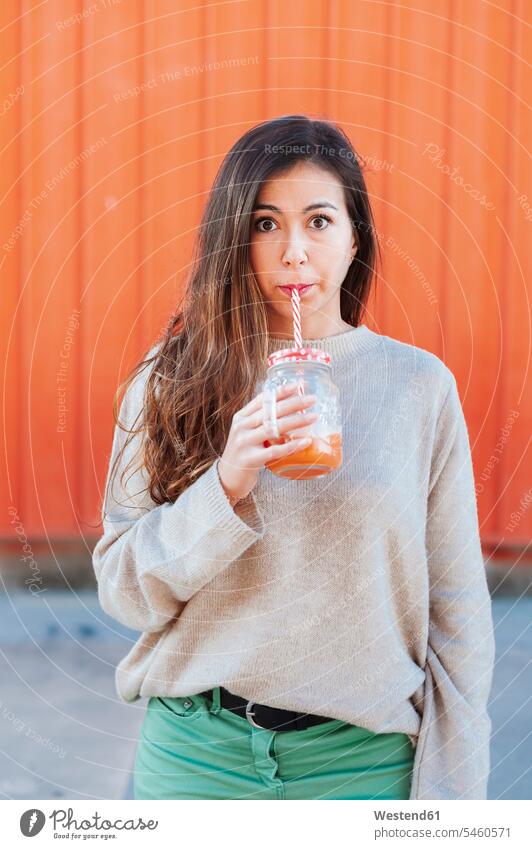 Schöne Frau trinkt Saft, während sie an einer Orangenwand steht Farbaufnahme Farbe Farbfoto Farbphoto Außenaufnahme außen draußen im Freien Tag
