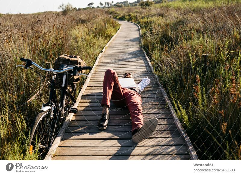 Gut gekleideter Mann mit Laptop liegt auf einem Holzsteg in der Landschaft neben einem Fahrrad Leute Menschen People Person Personen Europäisch Kaukasier