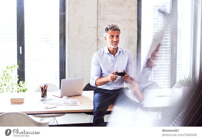 Lächelnder reifer Geschäftsmann sitzt im Büro auf dem Schreibtisch mit Controller Office Büros lächeln Portrait Porträts Portraits Businessmann Businessmänner