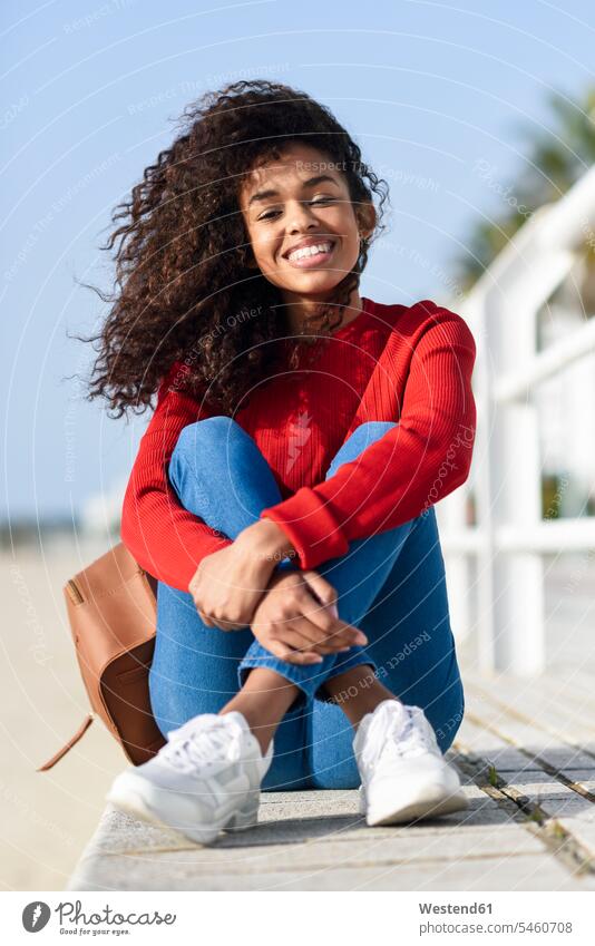 Porträt einer glücklichen jungen Frau, die auf einer Strandpromenade sitzt weiblich Frauen Strandpromenaden Beach Straende Strände Beaches sitzen sitzend