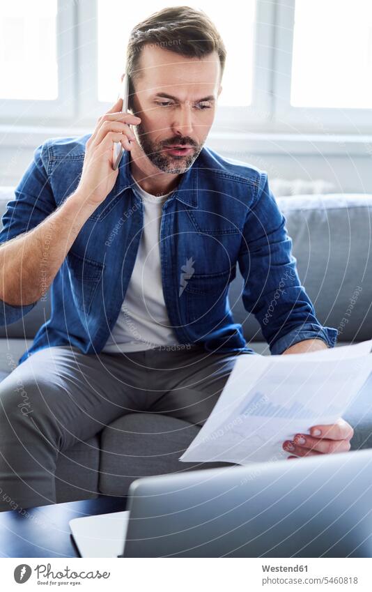 Ernster Mann mit Dokumenten und Laptop auf dem Sofa, der mit dem Handy telefoniert Couches Liege Sofas Männer männlich Notebook Laptops Notebooks telefonieren