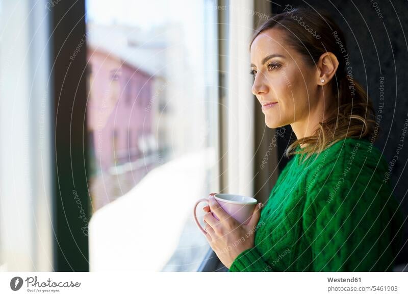 Frau hält Kaffeetasse, während sie zu Hause am Fenster steht Farbaufnahme Farbe Farbfoto Farbphoto Innenaufnahme Innenaufnahmen innen drinnen Tag