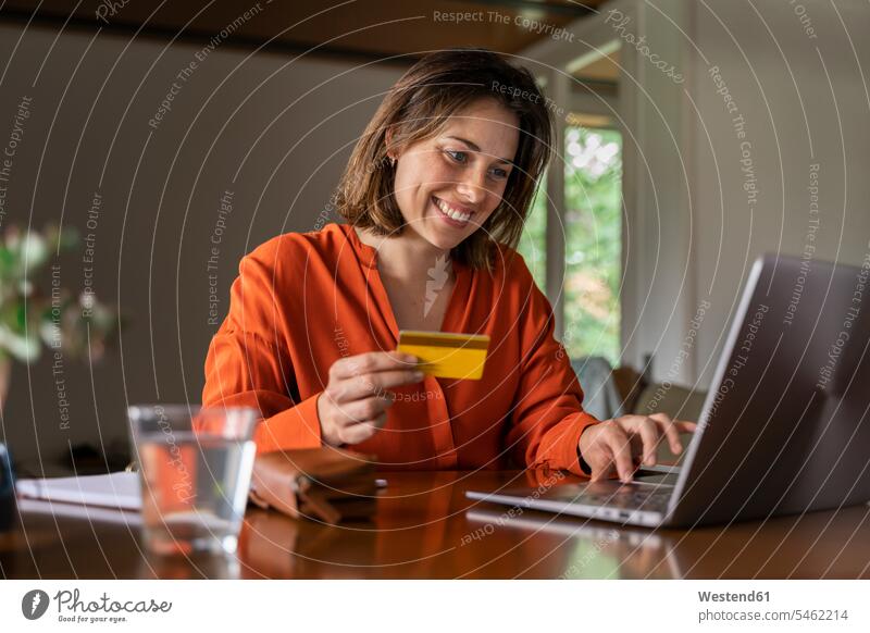 Lächelnde Geschäftsfrau mit Kreditkarte mit Laptop zu Hause Farbaufnahme Farbe Farbfoto Farbphoto Innenaufnahme Innenaufnahmen innen drinnen Tag