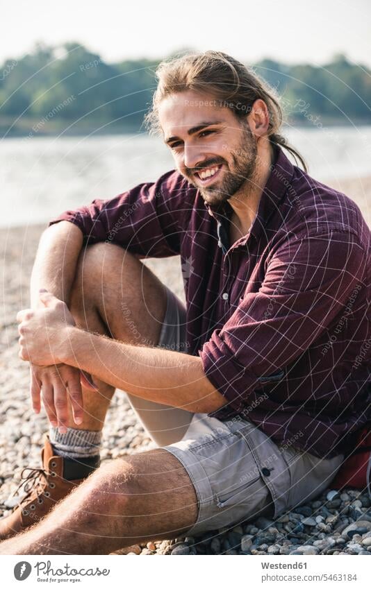 Lächelnder junger Mann sitzt am Flussufer Fluesse Fluß Flüsse lächeln sitzen sitzend Männer männlich Gewässer Wasser Erwachsener erwachsen Mensch Menschen Leute