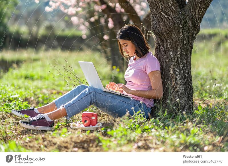 Teenager-Mädchen sitzt auf einer Wiese und lehnt sich mit einem Laptop an einen Baumstamm T-Shirts Rechner Laptops Notebook Notebooks Fotokamera Kamera Kameras