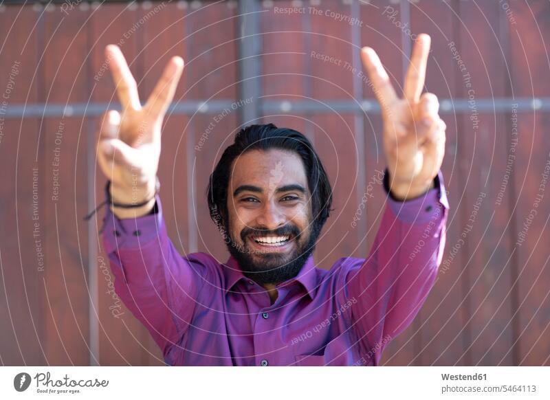 Junger lächelnder Mann macht Friedenszeichen vor einem Tor Job Berufe Berufstätigkeit Beschäftigung Jobs Techniken Technologie Techniker Hemden Arbeit