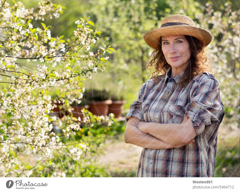 Porträt einer lächelnden reifen Bäuerin, die mit verschränkten Armen am Baum im Gemeinschaftsgarten steht Farbaufnahme Farbe Farbfoto Farbphoto Deutschland