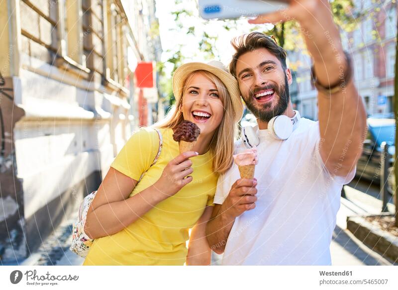 Glückliches junges Paar macht ein Selfie beim Eisessen in der Stadt Touristen Hüte T-Shirts Kopfhoerer Telekommunikation telefonieren Handies Handys
