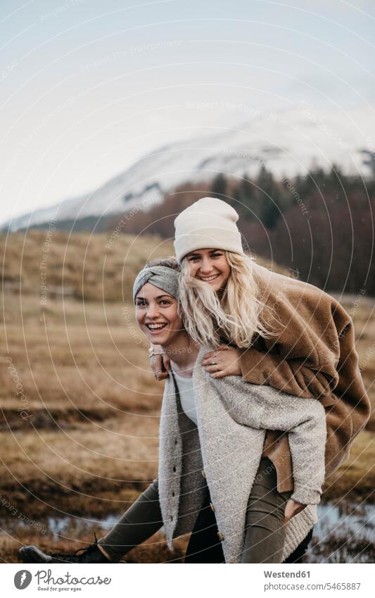 Großbritannien, Schottland, glückliche junge Frau trägt Freund Huckepack in ländlicher Landschaft Freundinnen Portrait Porträts Portraits tragen transportieren