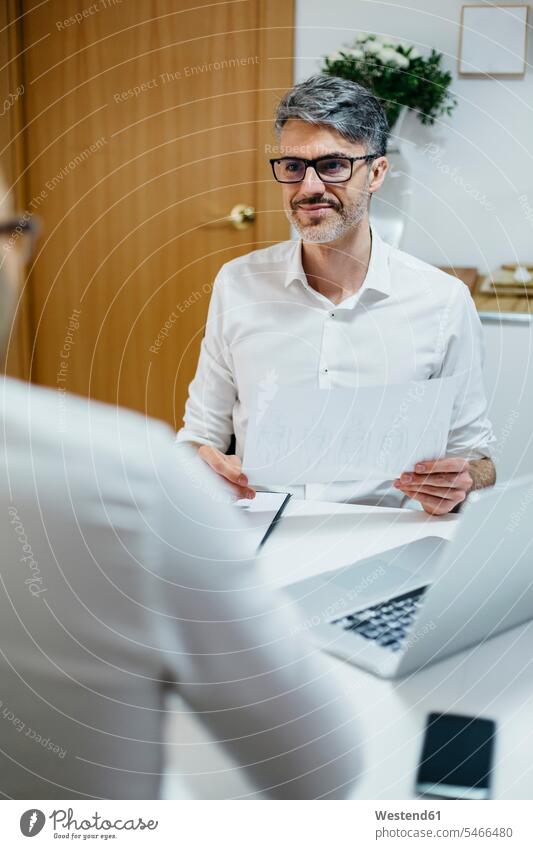 Lächelnder Mann am Schreibtisch mit Papier in der Hand schaut Kollege an Kollegen Arbeitskollegen Männer männlich ansehen lächeln Zuhause zu Hause daheim