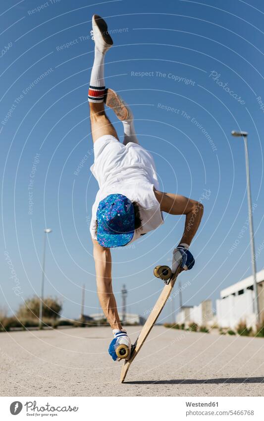 Rückenansicht eines Mannes in stilvollem, sportlichem Outfit, der auf einem Skateboard kopfüber vor blauem Himmel steht Trick skateboarden Rückansicht
